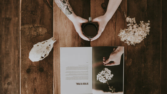 Hands holding a mug beside an open book and dried flowers on wood