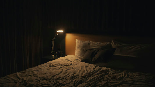 Dark bedroom at night, small bedside lamp glowing beside rumpled bed