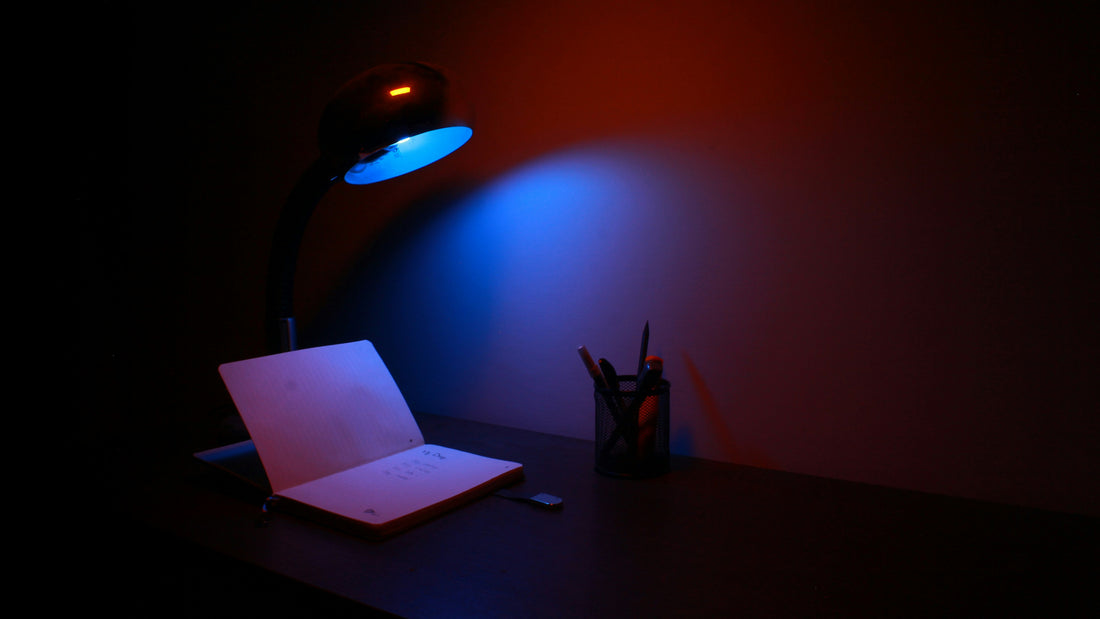 Desk lamp glowing over an open notebook and pencils in a dark room